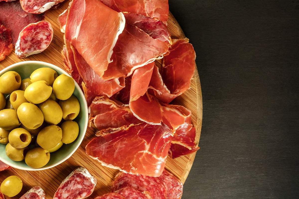 An overhead photo of a Spanish cold meats platter with jamon and payes sausage, with green olives, on a dark background.
