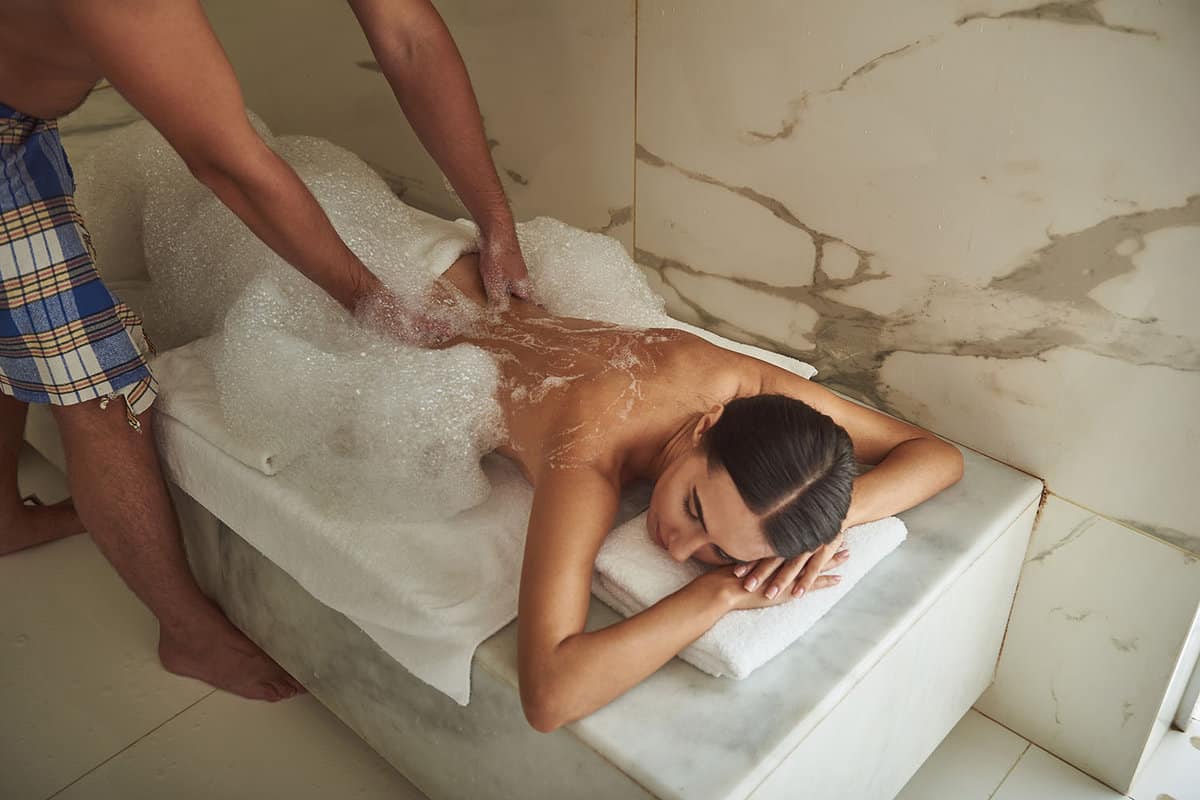Professional hamam worker touching the back of young lady while covering her with soap foam