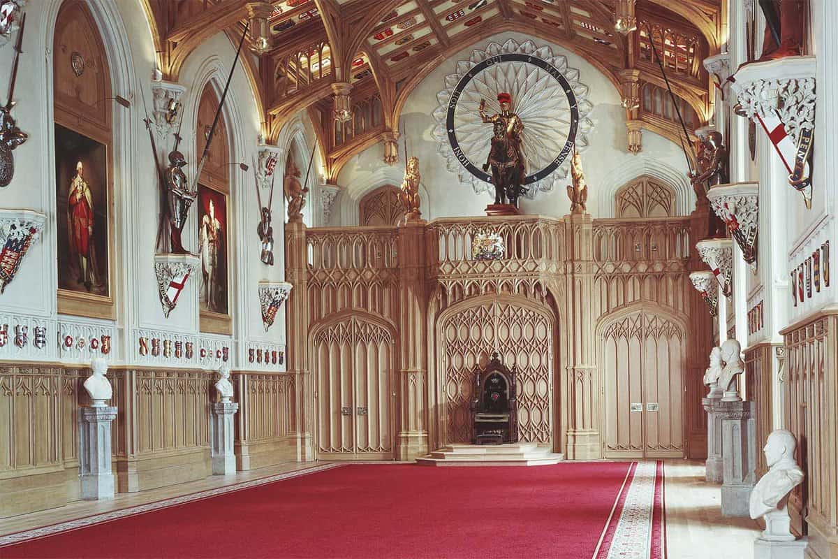 An inside look of St George's Hall in Windsor Castle. There is a contrast between the white, red and golden coloured interior with a red carpet laid in the middle of the room.