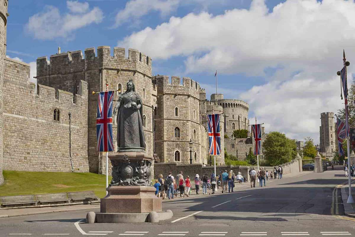 A side view of Windsor Castle that can be seen in the background. The Queen Victoria Statue is in the foreground with onlooking tourists