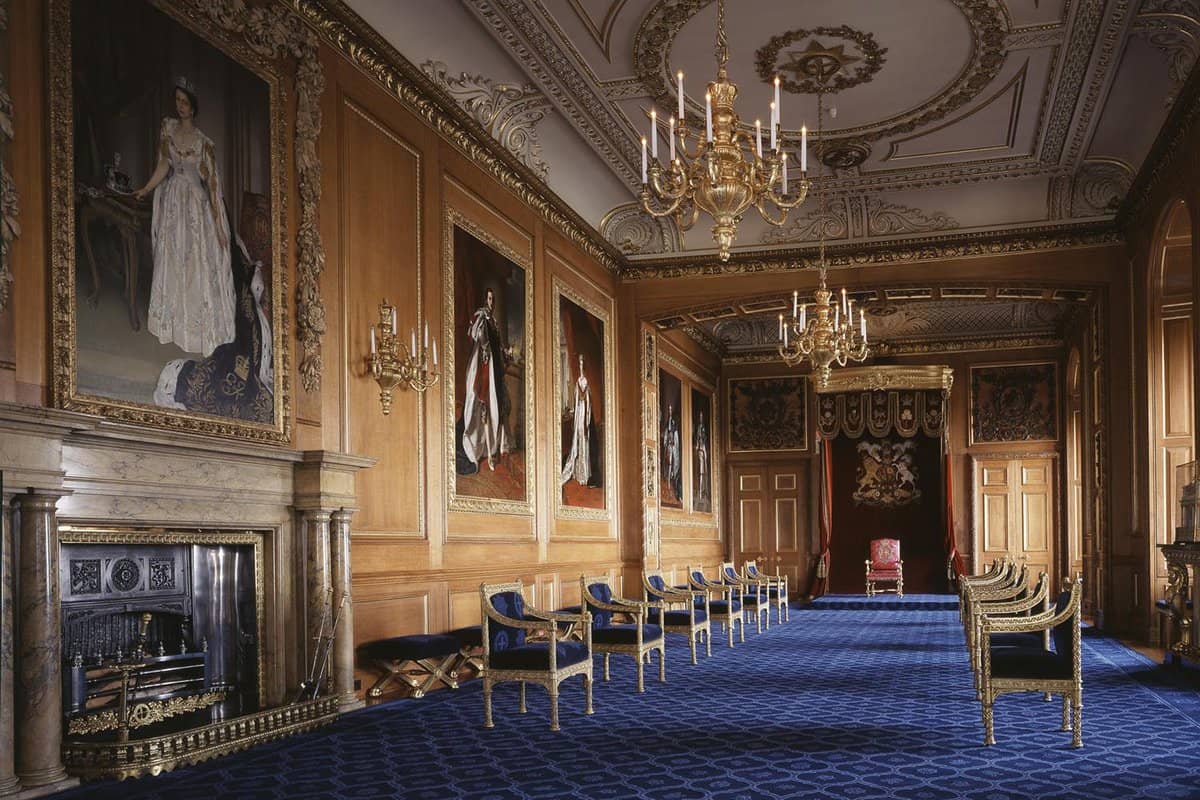 Inside Windsor Castle is the Throne Room that has wooden light brown walls and blue patterned carpet. There are multiple large paintings hung up on the walls with one above a fireplace. The room is lined with chairs along the side of the Throne room.