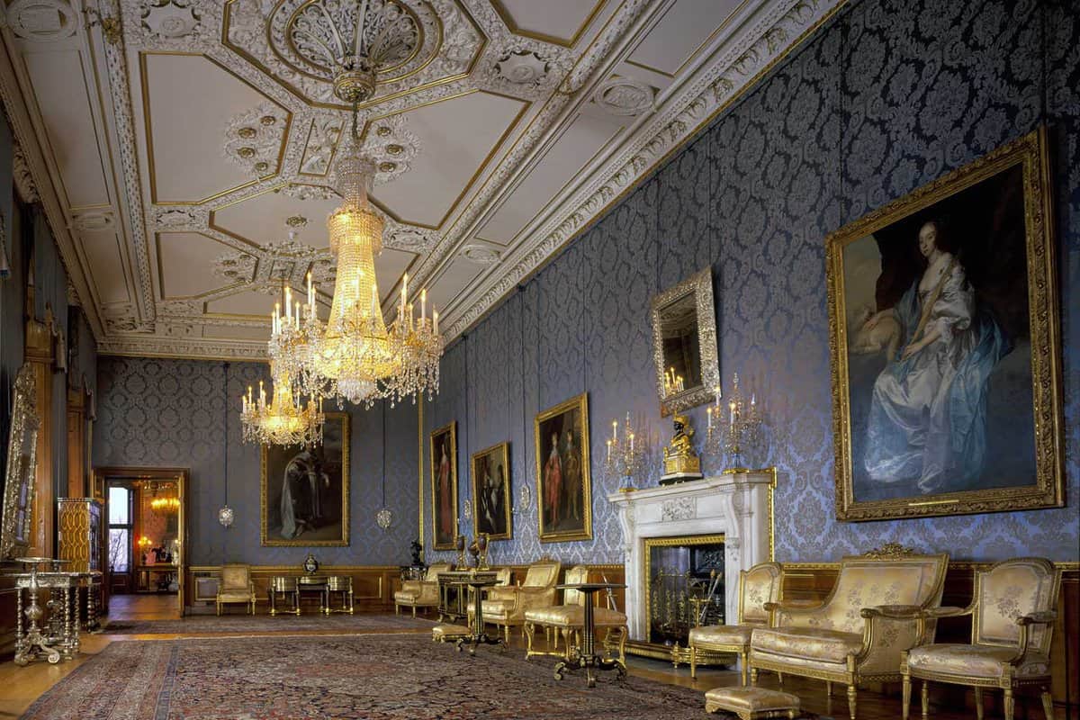 An inside view of the Queen's Ballroom in Windsor Castle. The floorspace has a large patterned carpet in the centre with two golden chandeliers. Alongside the room there are multiple gold coloured chairs with gold framed paintings hung on the walls.