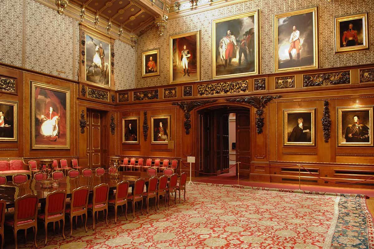Inside the Waterloo Chamber in Windsor Castle, there is a large polished brown table with a cluster of red chairs placed around the table. Around the walls there are several gold framed paintings that fill the wall up to the ceiling.