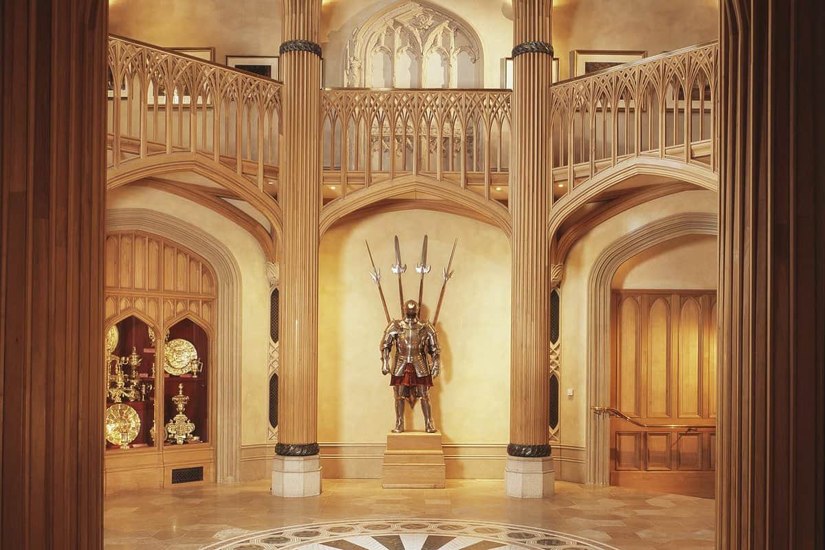 An inside view of the Lantern Lobby at the Royal Collection Trust. In the centre there is an armoured suit with four swords behind. Above this, there is an upstairs balcony curved around the building. On the left side, there are gold awards and medals showcased in the cabinet.