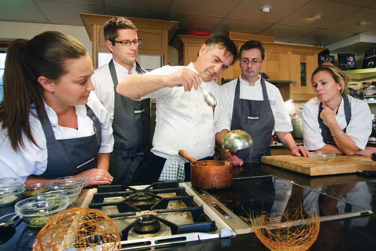 4 trainne chefs looking at Raymond Blanc whilst pouring some sauce`