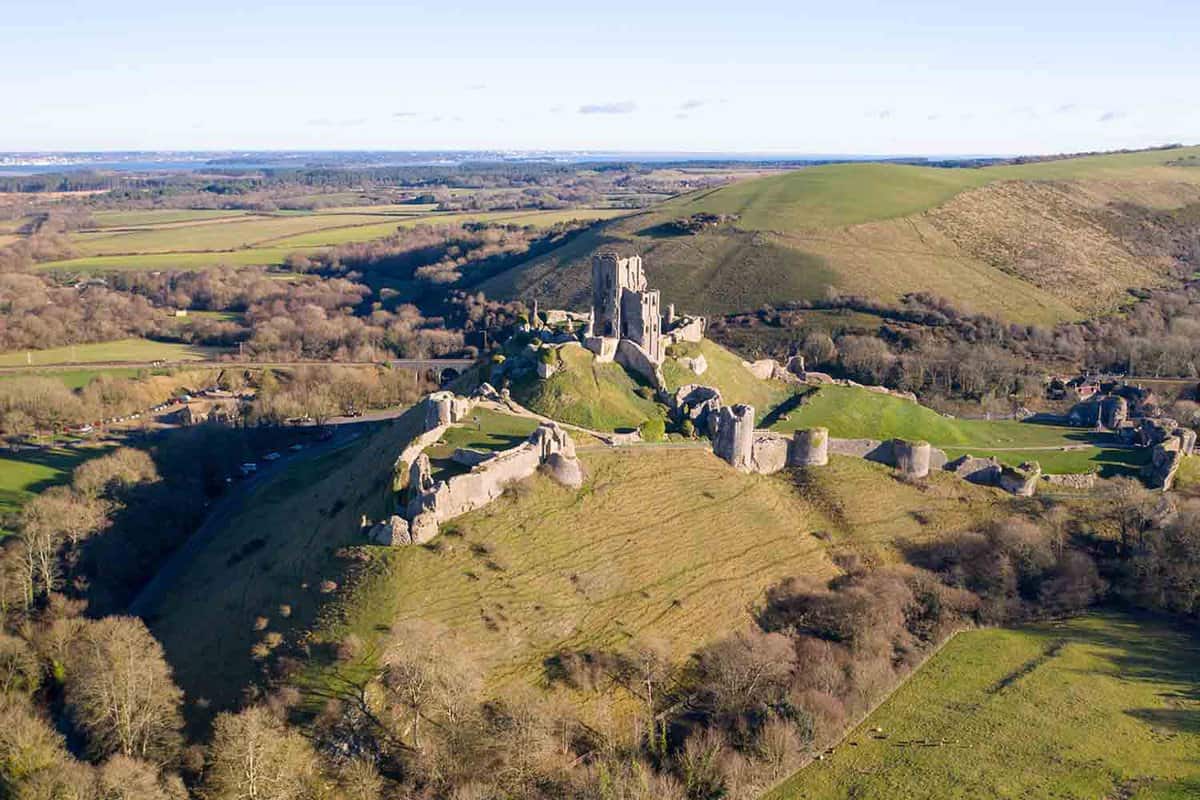 An aerial view of Corfe Castle situated on top of a hill with tress surrounding the bottom of the hill. In the background there is a large hill next to an open grass field.
