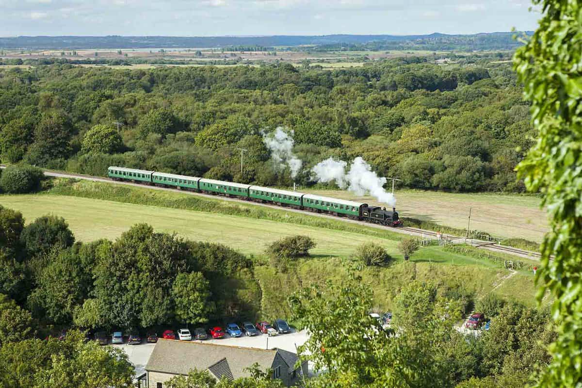 A view of a steam train from Corfe Castle on the Swanage railway line, surrounded by bright green trees and grass.