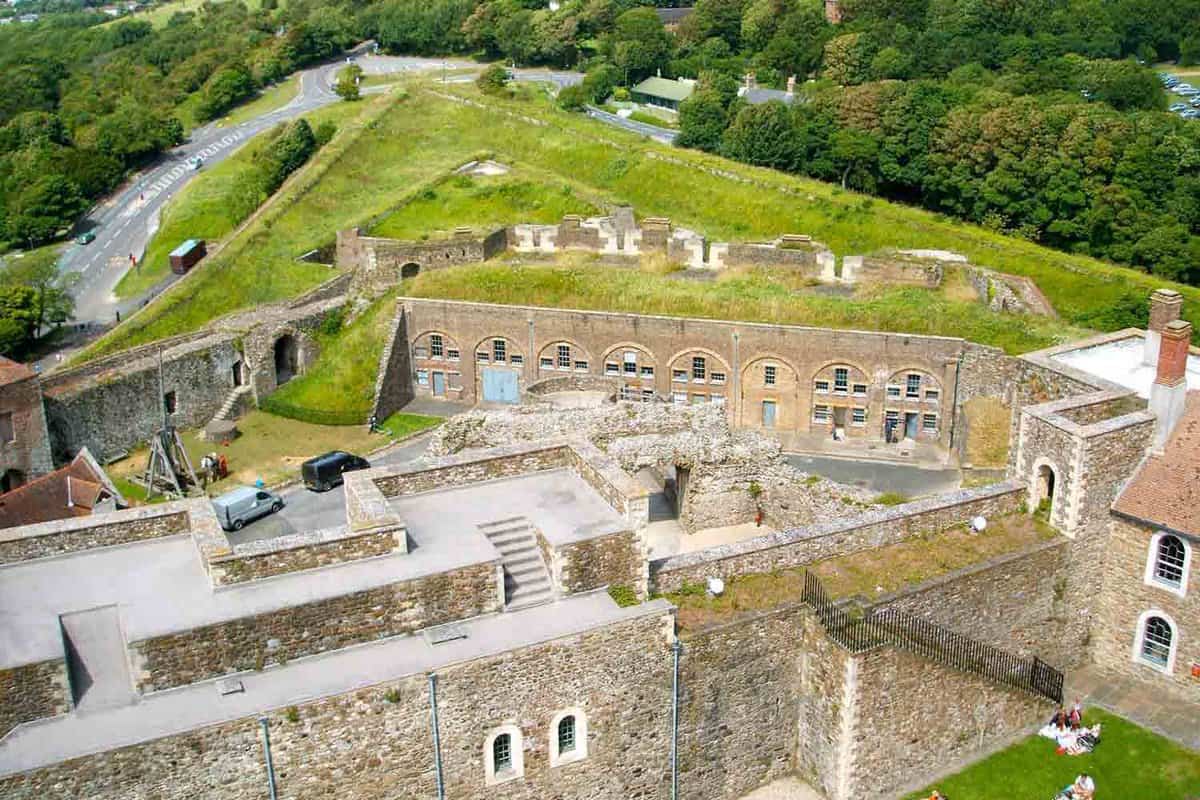 An aerial view of Dover Castle that displays an open and empty rooftop. In the background there is a road transport that is surrounded by the woodlands.
