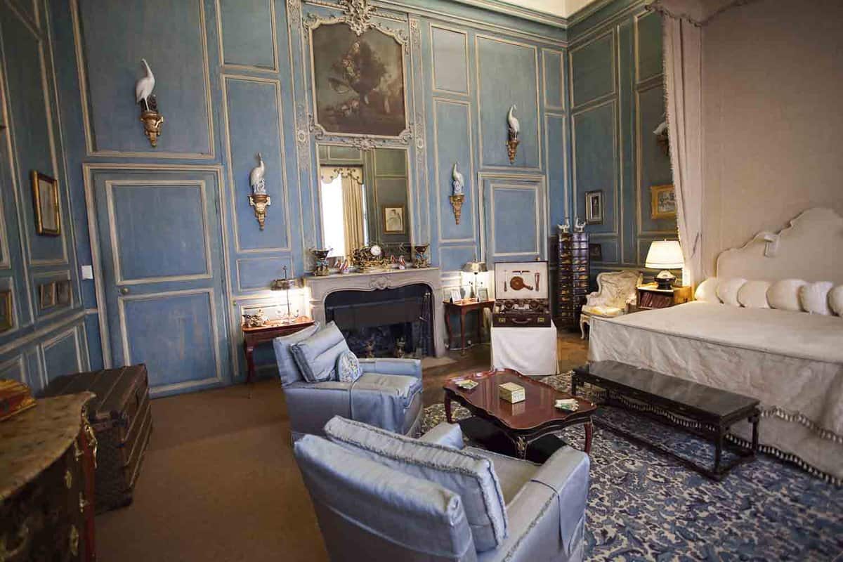 Interior view of typical bedroom in an English castle. Two single chairs around a round table in the centre of the room. A mirror on top of the fireplace with wood furniture placed around the room