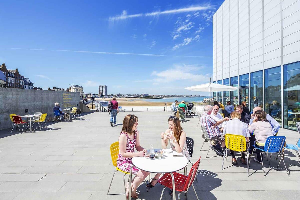 An outside view of an open seating area right beside Turner Contemporary art gallery. In the foreground there are a few people seated on chairs with tables in groups. In the background the sand can be seen peeking through with a bright blue sky.