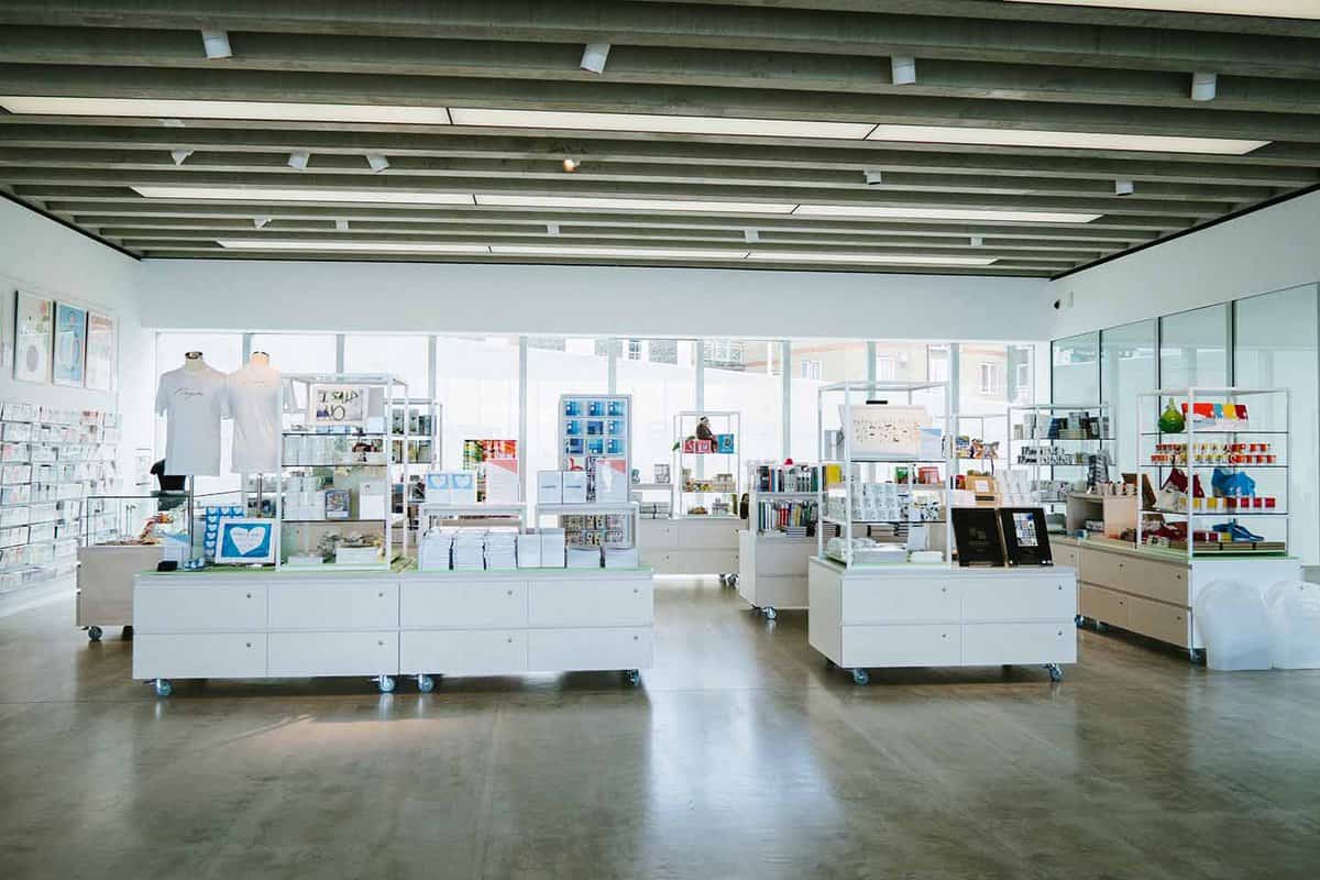 An inside view of Turner Contemporary art gallery shop with items and souvenirs related to the gallery. In the background there is a wide stretching set of windows and horizontal lined lights on the ceiling, giving the room a bright atmosphere.
