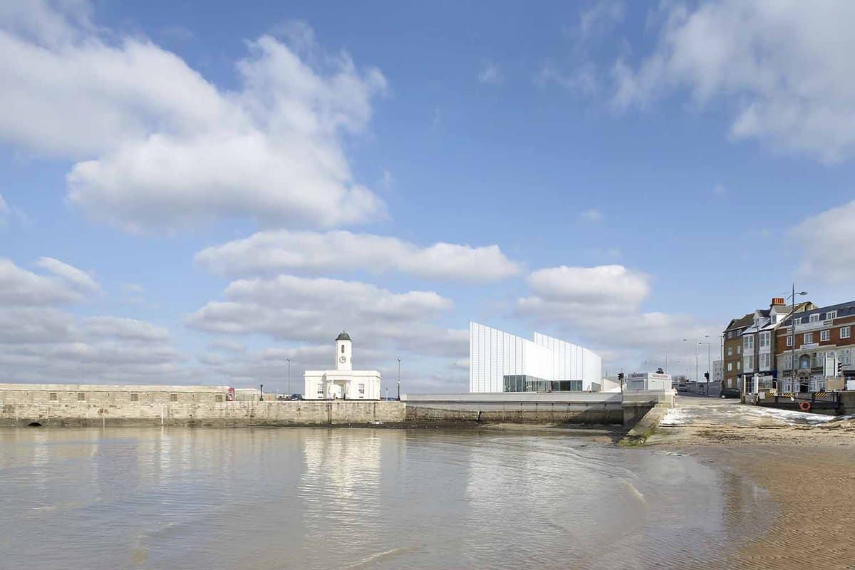 A landscape view of Turner Contemporary art gallery in Margate. The foreground shows the sandy beach of margate against the tides, where the gallery can be seen in the distance and reflected on the surface of the waters.