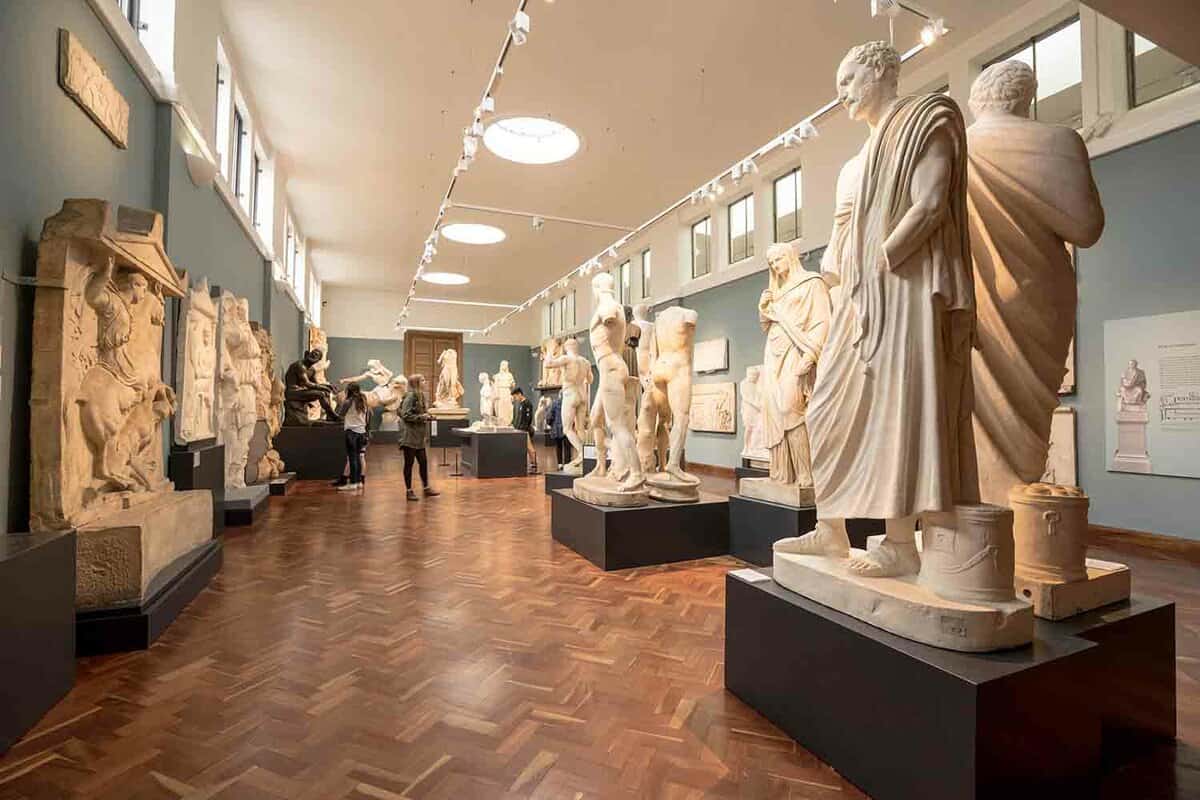 Several marble carved statues of figures in sweeping robes. They are positioned on plinths within a viewing gallery, with a few visitors looking at them.
