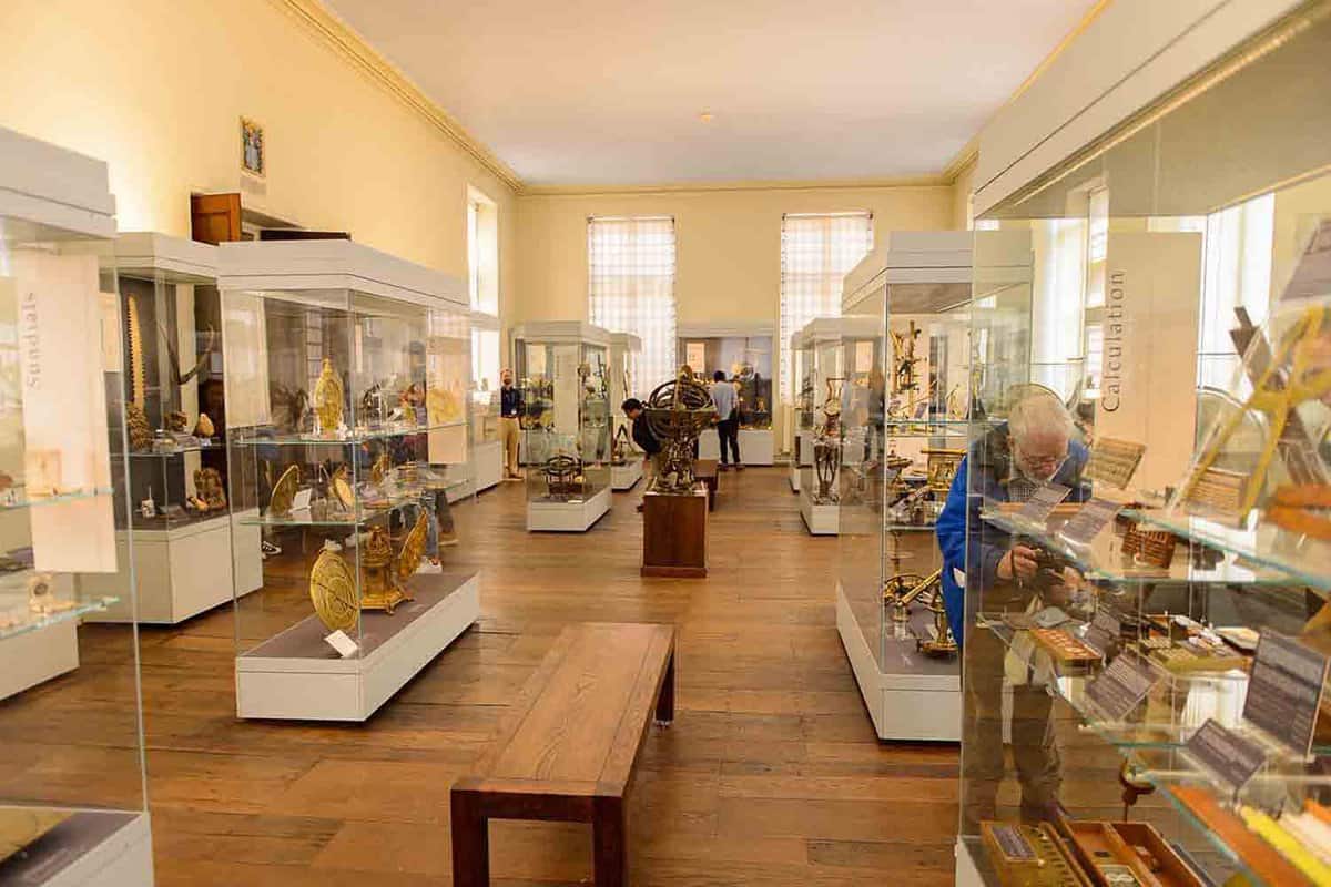 A collection of scientific instruments from Middle Ages to the 19th century, displayed in glass cabinets. There is a visitor peering intently at one cabinet, and a bench in the centre of the room.