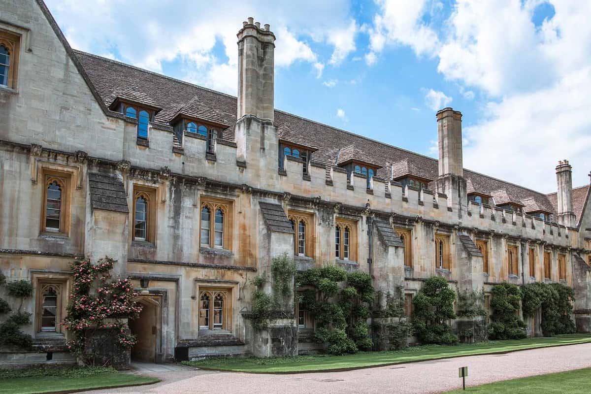 The inner yard and campus buildings of Magdalen College. A long pale grey sandstone building, with three chimneys rising at regular intervals. The building is low to the ground with many square windows with pale wood window frames. There is green foliage in rough rectangle shapes climbing the façade at regular intervals. An arched doorway leads inside the building, and there is a pathway leading past the building with grass on either side.
