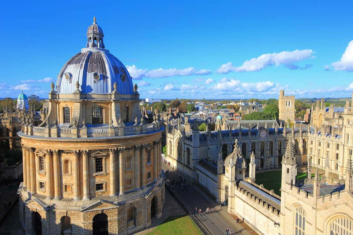 Below a bright blue sky, All Souls College sits to the right, with the Radcliffe Camera to the left. The buildings are neo-classical in style, with a road running between them.
