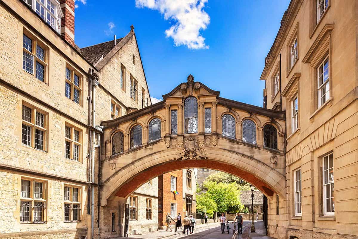 The Bridge of Sighs (Hertford College) on a sunny day. The bridge runs between two buildings above a cobblestone street underneath which people are walking. The bridge is arched sandstone with rectangular glass windows. The street is illuminated by afternoon sun.