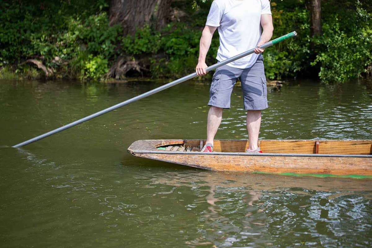 A close up of a person standing up and pushing a punt along using a pole. He is wearing a white t-shirt and grey shorts, and is holding the punting pole with both hands. The end of the pole is in the water, which is a grey-green colour.