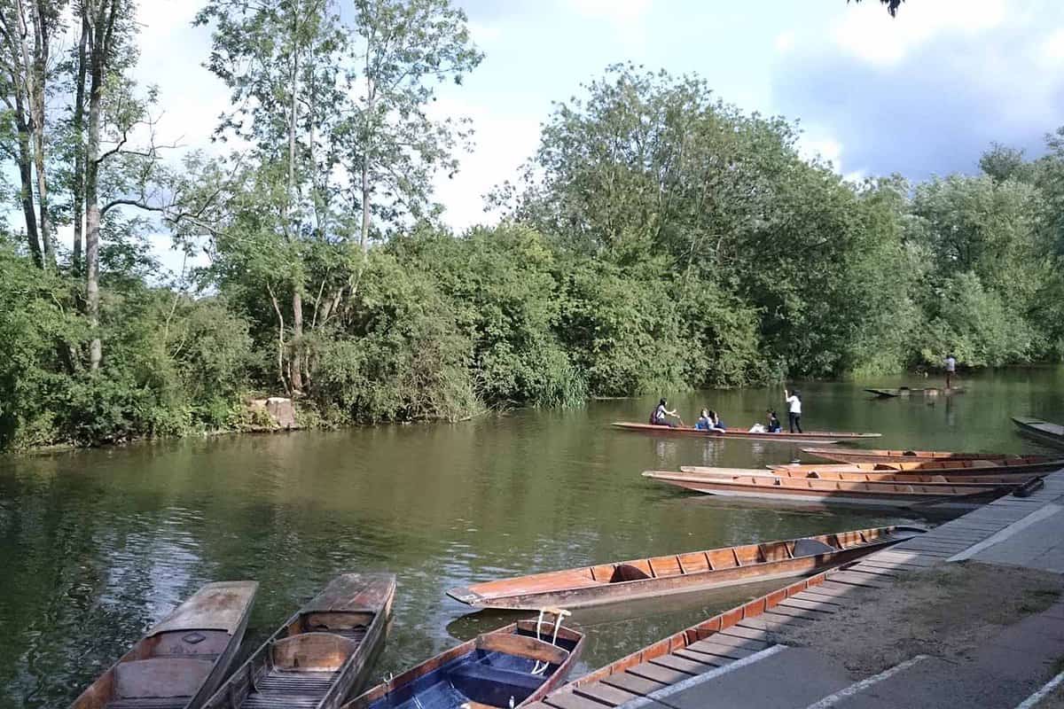 A view from the riverbank punt station, where there is a restaurant (not pictured). The opposite riverbank has green foliage lining it. In the foreground there are several moored punts. The photo focuses on a figure pushing a punt along down the river.