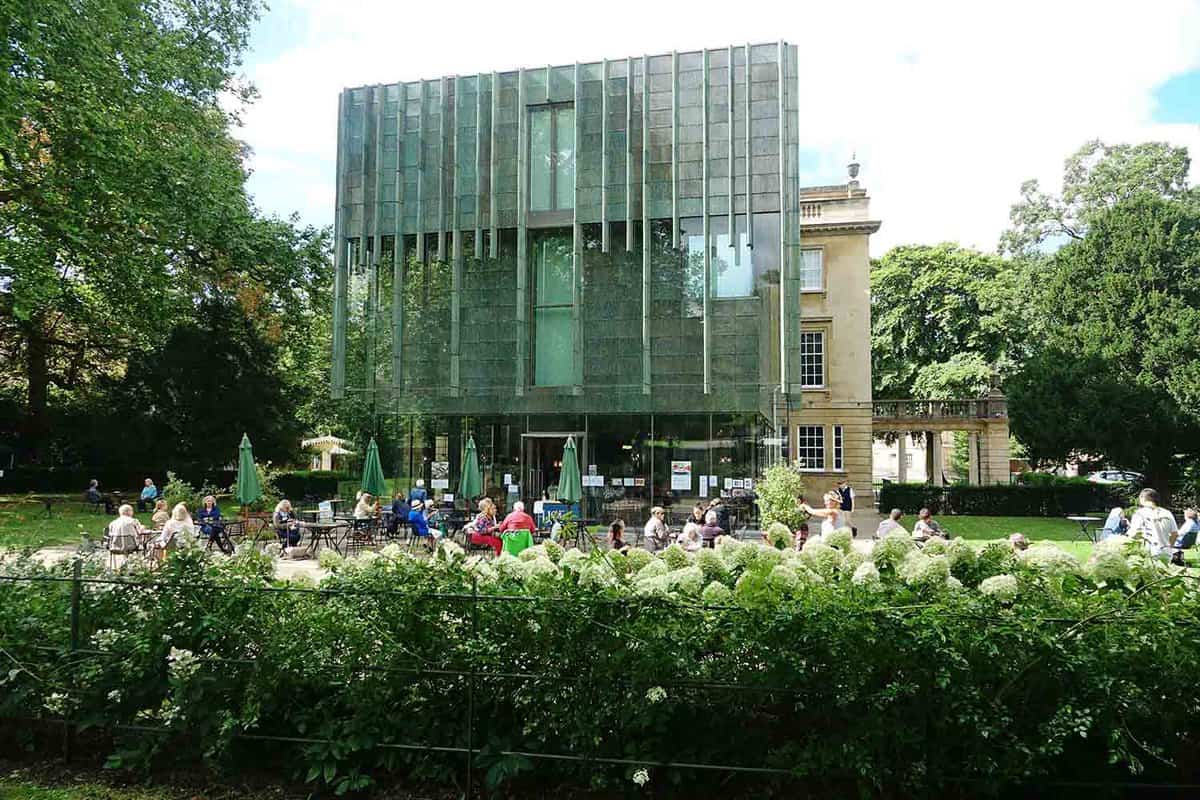 The modern glass add on to the Grade 1 listed building as seen over a hedge of flowers. People at cafe tables.