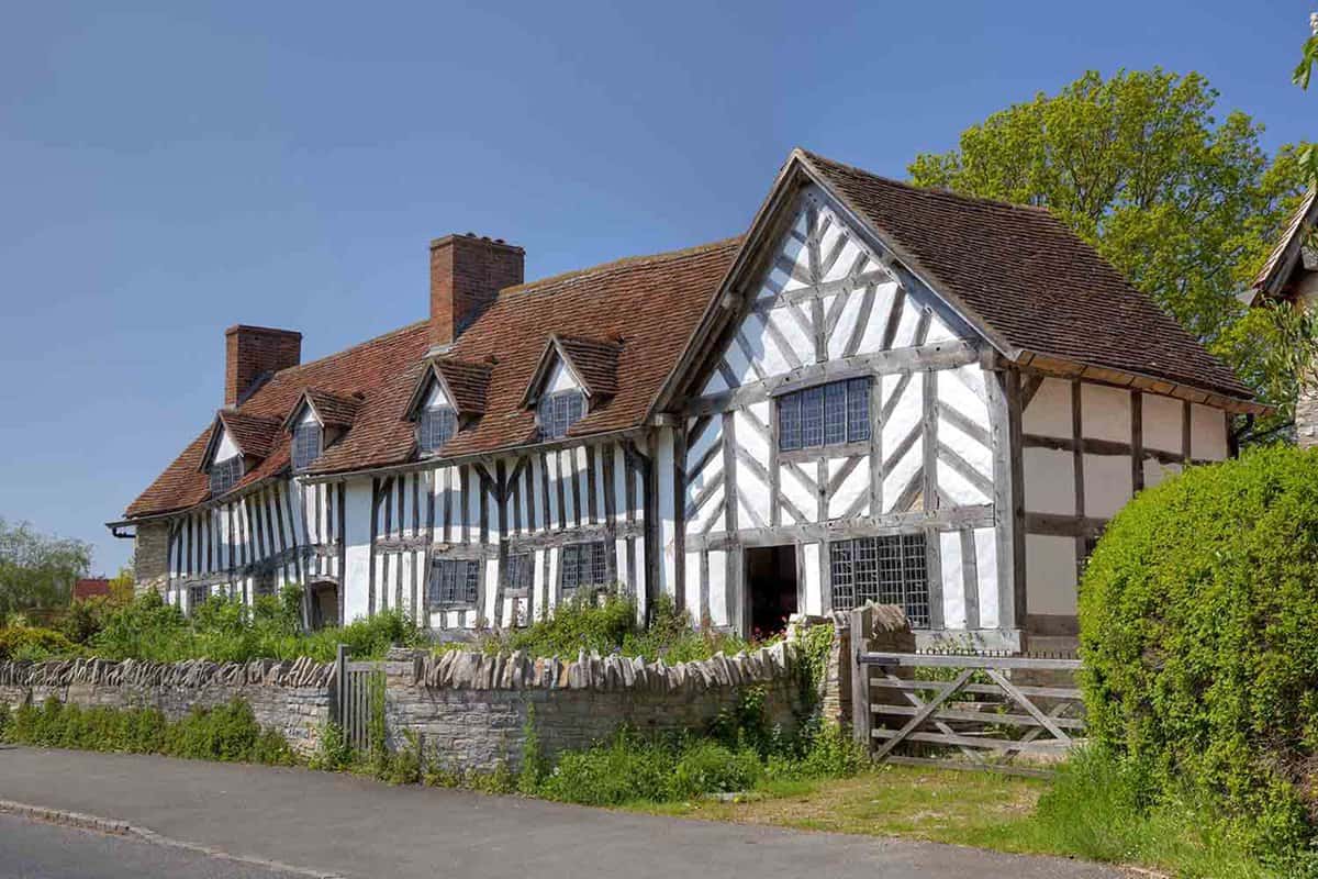 An outside view of Mary Arden's House (William Shakespeare's Mother). A large white farmhouse that has brown roofing and accents with a brick entrance and a wooden gate.