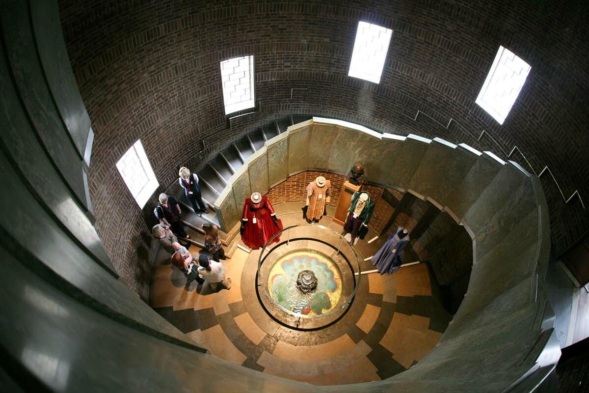 A photo of a small old fashioned curved stairwell with a small fountain at the centre, with ongoing visitors looking at the fountain