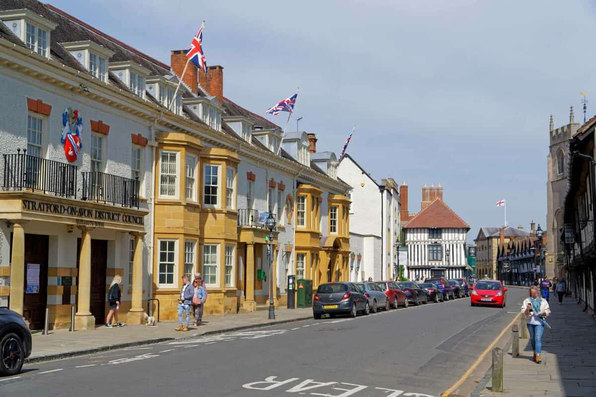A side view of Stratford-on-Avon District Council, with three United Kingdom flags mounted on the roof. The building is painted in a bright yellow colour that stands out against the surrounding white buildings on this high street.