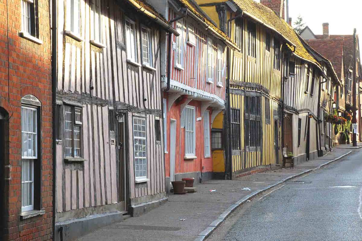 A row of medival houses along a small road. Each house is painted a different colour and on the picture there are pink, yellow, peach and brown houses. Along with a bricked modern house in the foreground that displays the change since Medieval times.