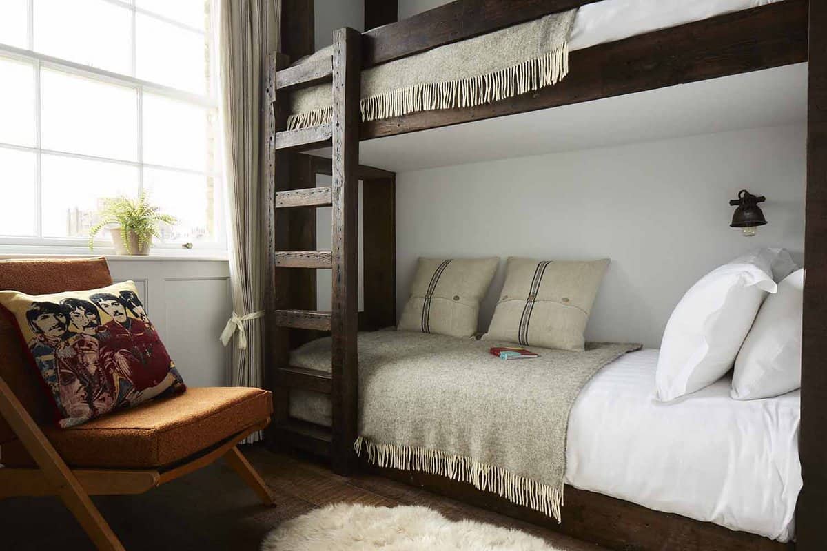 A bunk bed made of dark wood, made up with white sheets and a beige blanket and pillows. A cushioned chair sits next to the bunk beds, and a small plant sits on the windowsill.