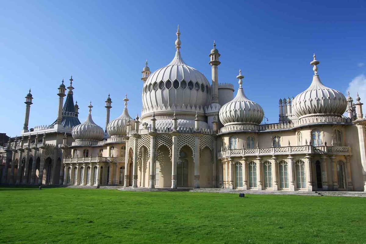 The Brighton Royal Pavilion. A building in Indo-Saracenic style, with domes and minarets. It is made of pale stone and sits atop green grass, under a bright blue sky.