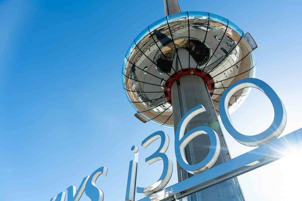 A shot looking upwards at the Brighton i360. A 162m viewing tower that is pole shaped, with the viewing area shaped like a donut around the central pole. The i360 sign is blue and seen from below, and the tower is white against the blue sky.