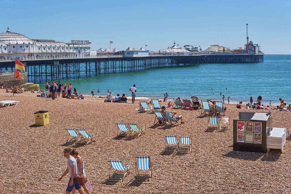 Brighton beachfront on a sunny day. Beach chairs are on the shingle in pairs, and beachgoers are walking or sitting near the shore. In the background to the left, the pier just out into the sea. You can see the fair rides at the end of the pier interrupting the horizon.