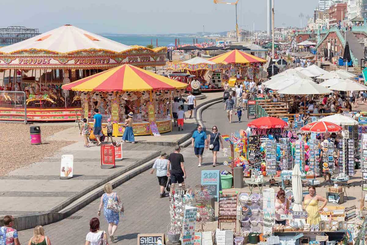 A street scene on Brighton seafront in the summer. There are souvenir shops and children's rides, and it is full of tourists enjoying their visits. There are shopping stands, and people walk along the boardwalk. The sea is on the horizon in the background.