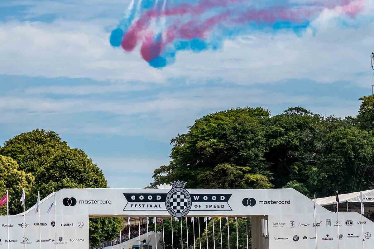 Crowds of spectators watch as the Royal Airforce Aerobatic Team (Red Arrows) fly with blue and red smoke. A race car passes beneath the Festival of Speed bridge.