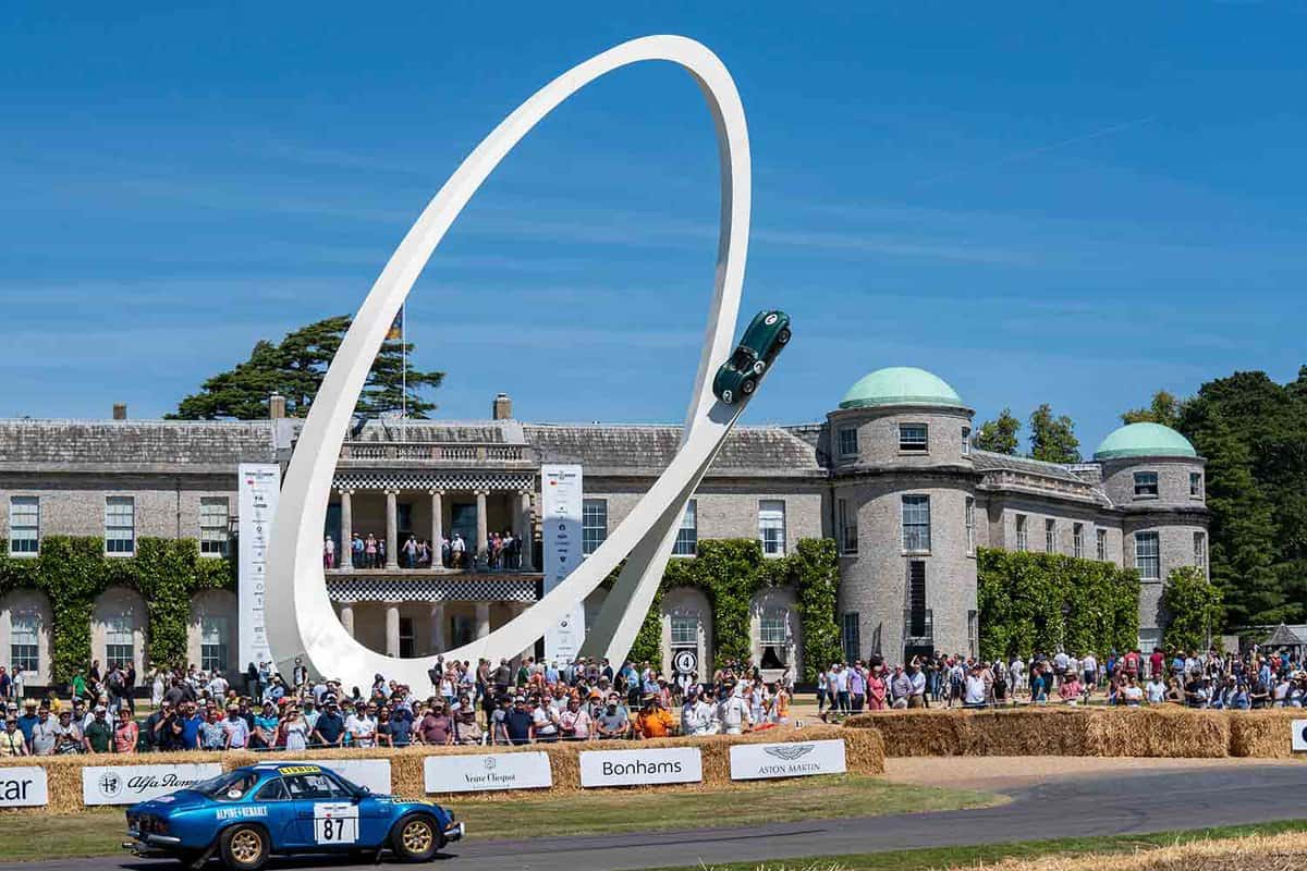 Crowds gather in front of Goodwood House on the first day of the Festival of Speed, a four day celebration of motor sport. A blue Renault race car flies past.