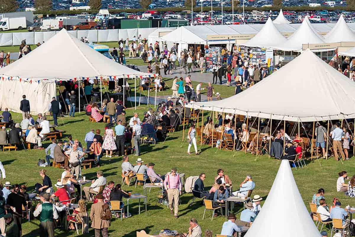 Looking down across white canvas tents covering shops, bars and cafes in a field at Revival. Crowds wearing vintage clothing sit and enjoy the sunshine.