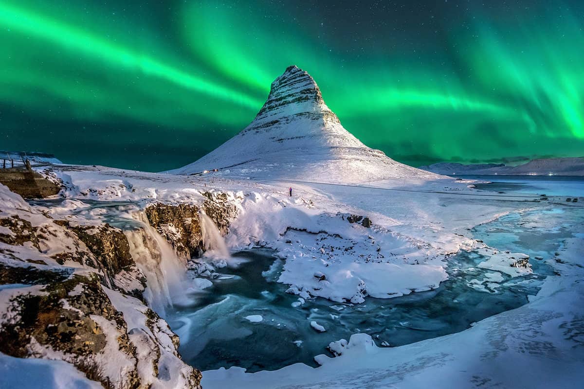 snowy landscape at night with a pointed mountain, and northern lights in sky behind it