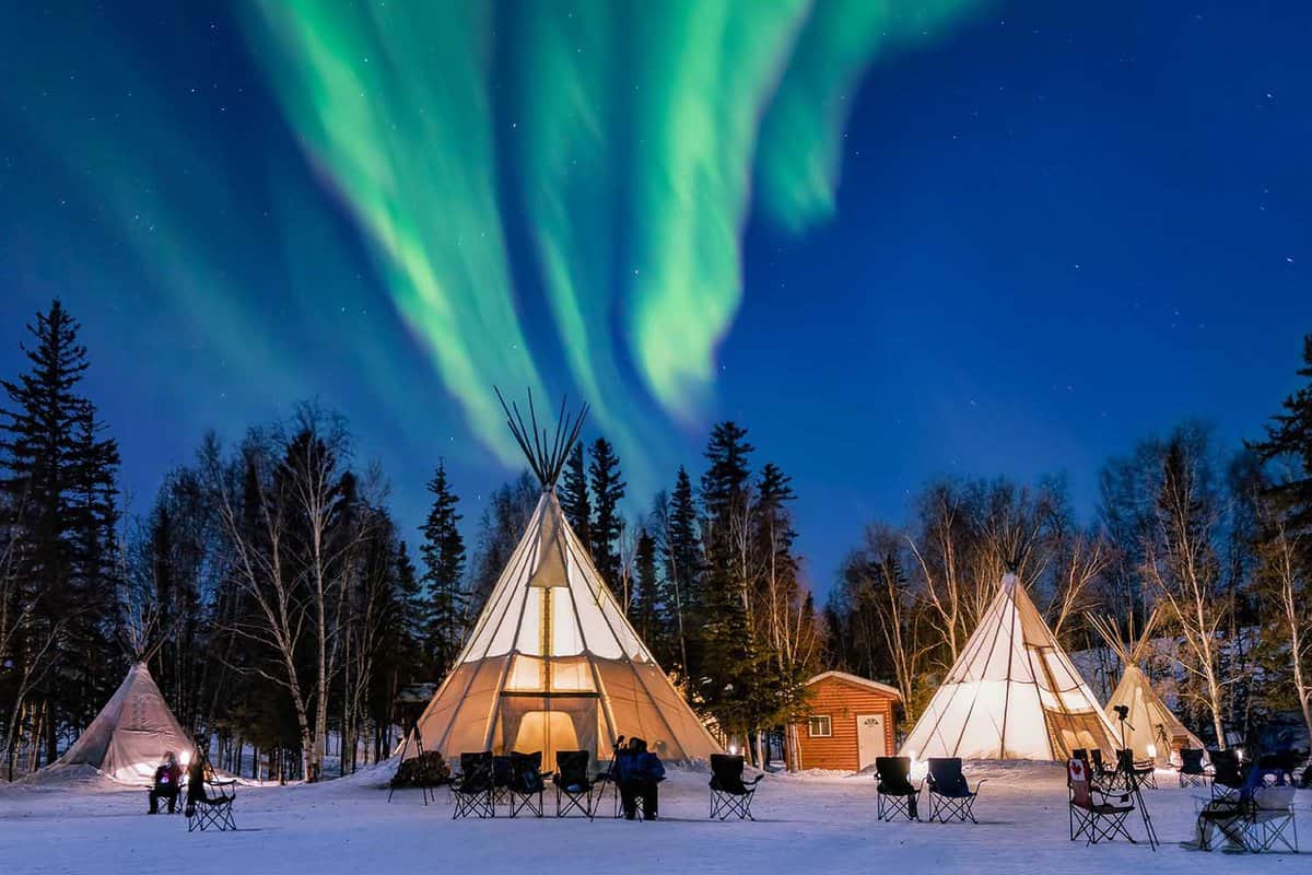Small tepee village with people sitting outside tents staring up at Northern Lights
