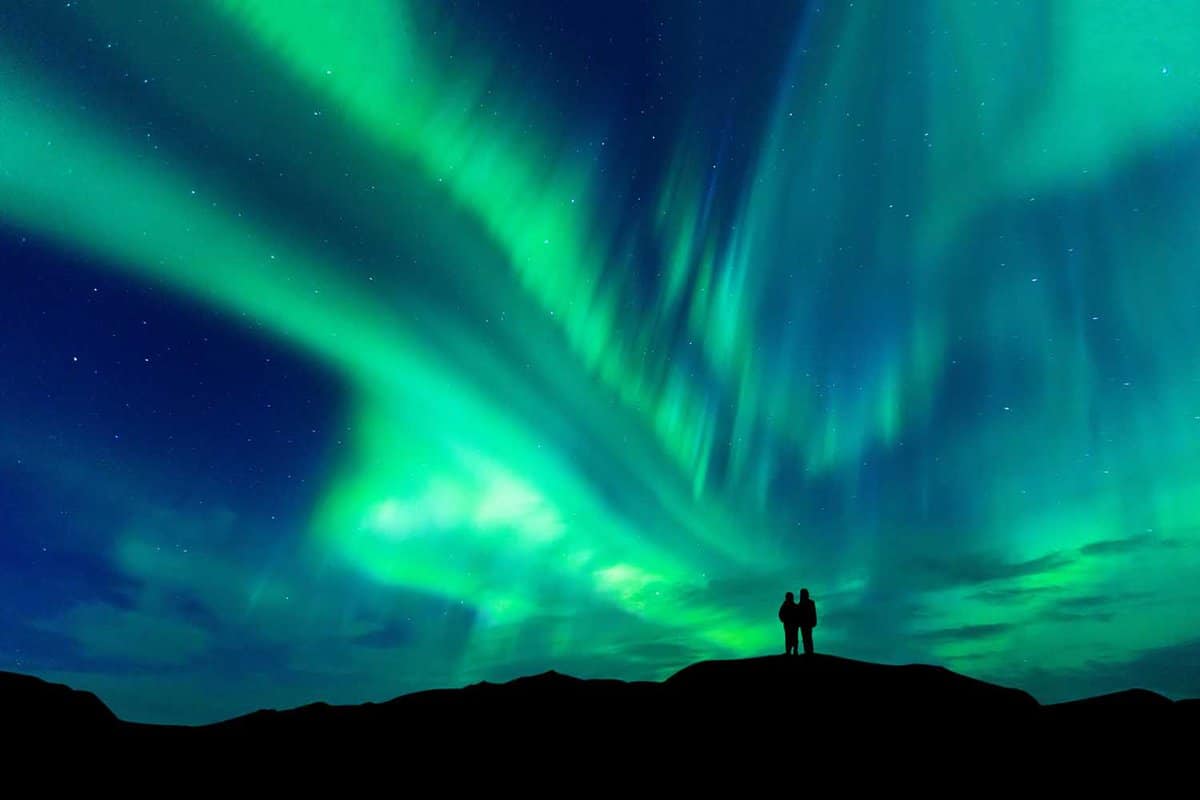 Dark silhouettes of a couple standing on hilltop with Northern Lights behind